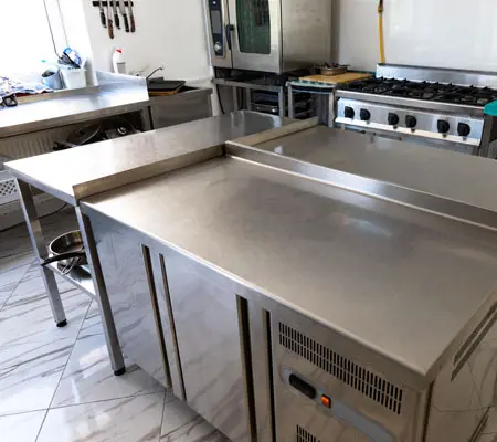 Construye paso a paso tu propia mesa de acero inoxidable ¡Es muy sencillo! 10 Conjunto de tres mesas de acero inoxidable al centro de una cocina de restaurante, con una estufa y horno industrial al fondo