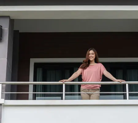 Barandas para Escaleras de Acero Inoxidable ¡Constrúyelas fácilmente! 4 Mujer con blusa rosa y pantalón beige en la orilla de un balcón con baranda de acero inoxidable viendo al exterior, al fondo se ve el ventanal de un departamento