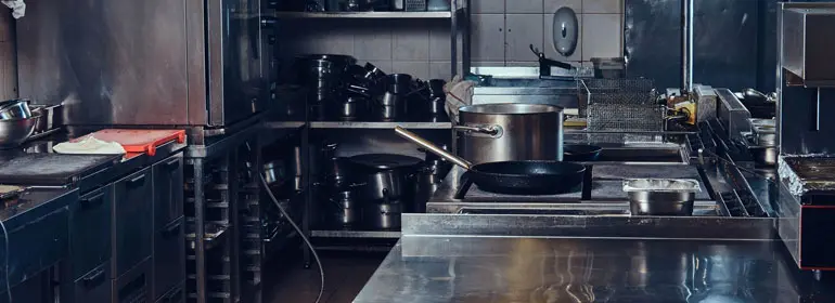 ¿Cómo elegir muebles de acero inoxidable? ¡Descubre sus beneficios! 1 Primer plano de una cocina con muebles de acero inoxidable, del lado derecho está una mesa y una freidora, al fondo un anaquel y de lado izquierdo un refrigerador y estantería