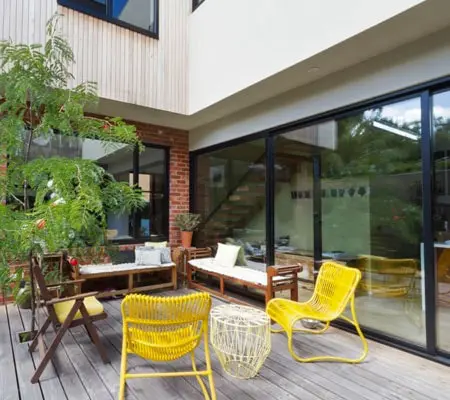 Puertas de Aluminio ¡La mejor opción para el hogar! 2 Vista lateral del patio de una casa con muebles para exterior y un árbol mediano del lado izquierdo, al fondo se ve una amplia puerta junto al resto de la casa de dos plantas