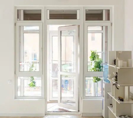 Puertas de Aluminio ¡La mejor opción para el hogar! 1 Vista frontal de una ventana color blanco y vidrio, se encuentra en un cuarto que da a un balcón, del lado derecho hay un mueble de madera con varias repisas.