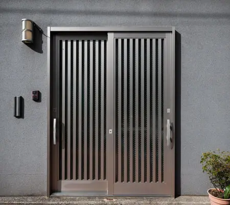Puertas de Aluminio ¡La mejor opción para el hogar! 5 Vista frontal de una puerta de aluminio corrediza color gris metálico ubicada al frente de una pequeña casa color gris, a la derecha hay pequeñas macetas decorativas