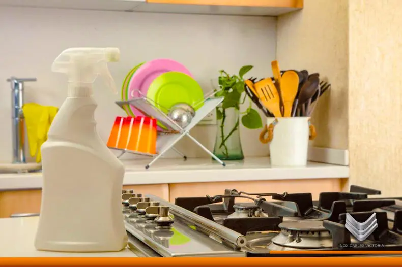 Estufa de acero inoxidable, a un lado hay un producto para su limpieza y al fondo se observa un trastero y diversos elementos de decoración de una cocina