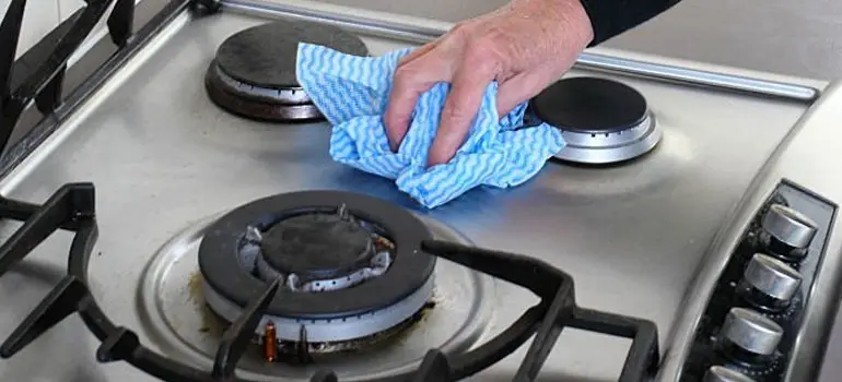 Pasos para limpiar Estufa de Acero Inoxidable 1 Manos de una persona con blusa negra limpiando una estufa de acero inoxidable con un paño azul
